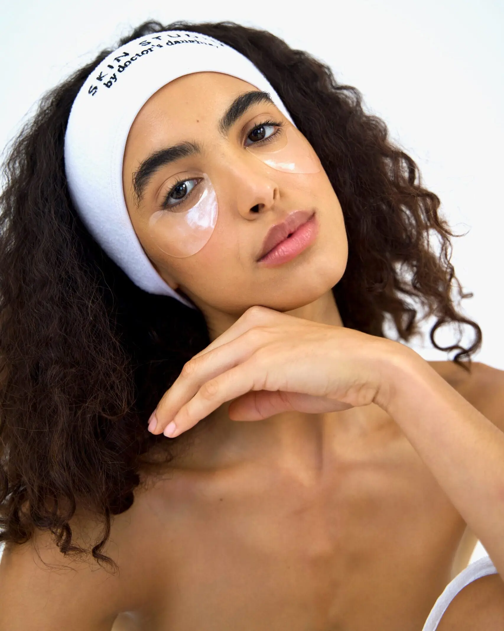 Woman with glowing skin wearing under-eye patches and a "Skin Studio by Doctor's Daughter" headband, highlighting a fresh and relaxed appearance