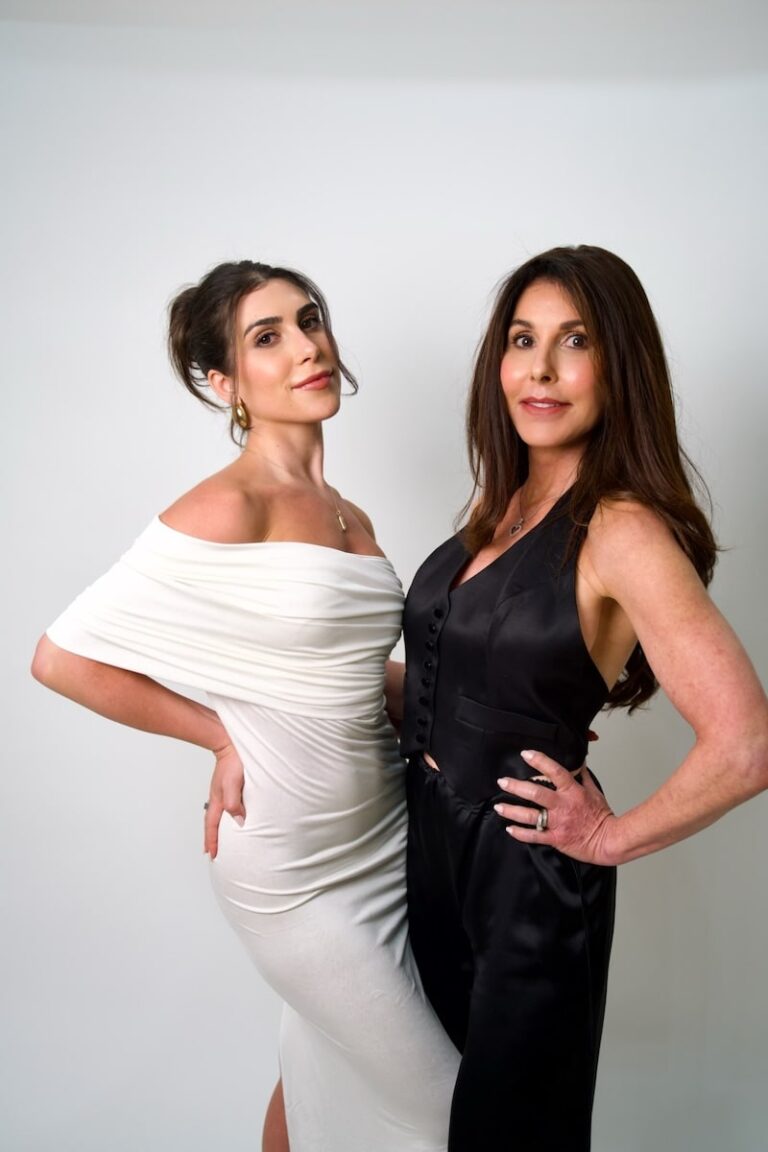 Two elegant women in white and black dresses posing confidently against a plain backdrop.
