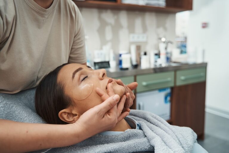 Woman with under-eye patches receiving a facial massage at a spa.