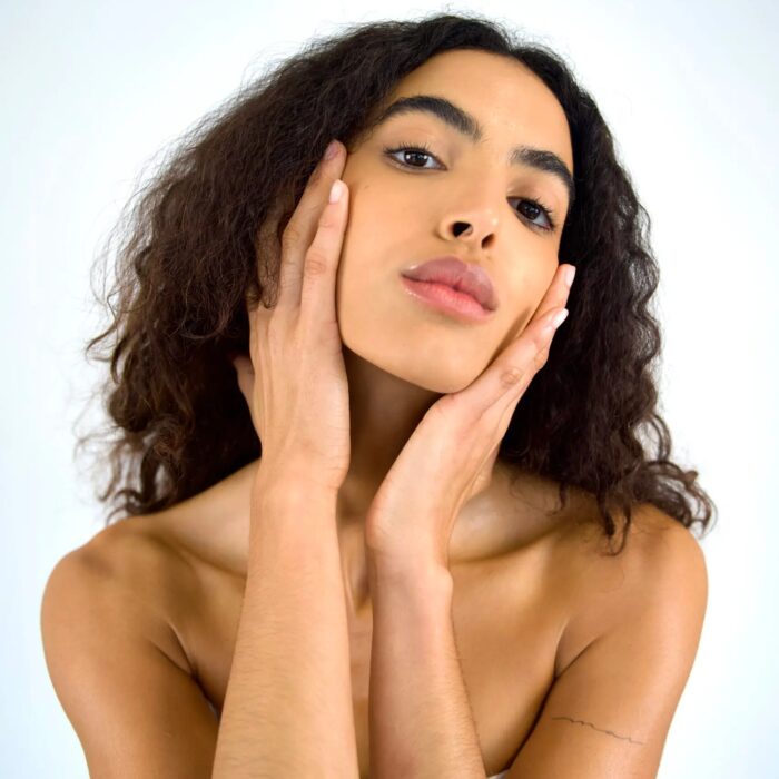 Woman with curly hair posing with hands on her face against a soft background.