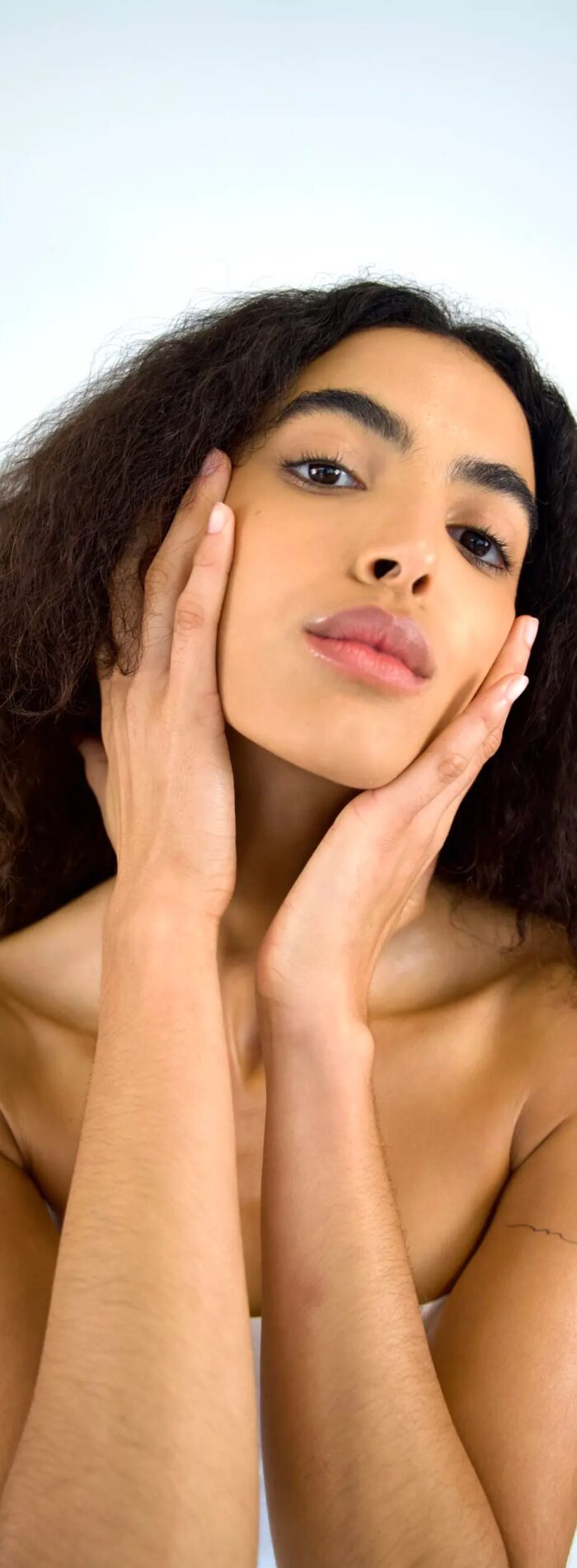 Woman with curly hair posing with hands on her face against a soft background.